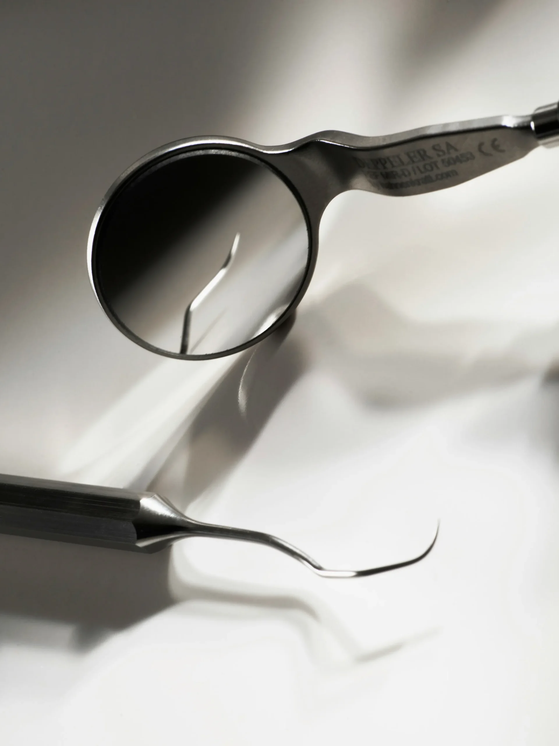 Primer plano de un espejo intraoral clínico de la marca Deppeler y una sonda dental de exploración metálica sobre una superficie blanca iluminada en la clínica Dentelier en Alicante.
